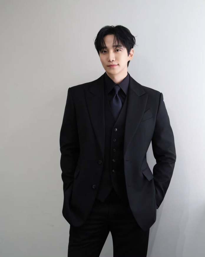 A man in a black suit with a fitted vest and tie, standing confidently against a light gray wall.