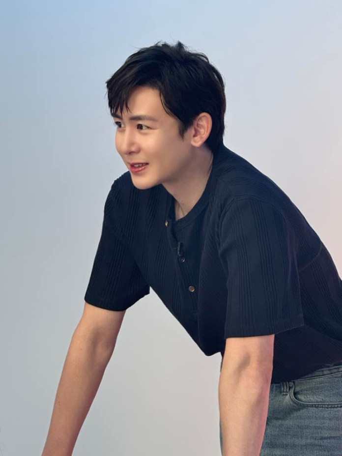A young man with dark hair smiling while leaning forward, wearing a black shirt and light jeans against a soft blue background.