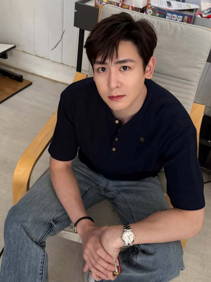 A young man with short dark hair sits on a chair, wearing a navy blue shirt and light blue jeans. He has a watch on his left wrist and is looking directly at the camera with a neutral expression.