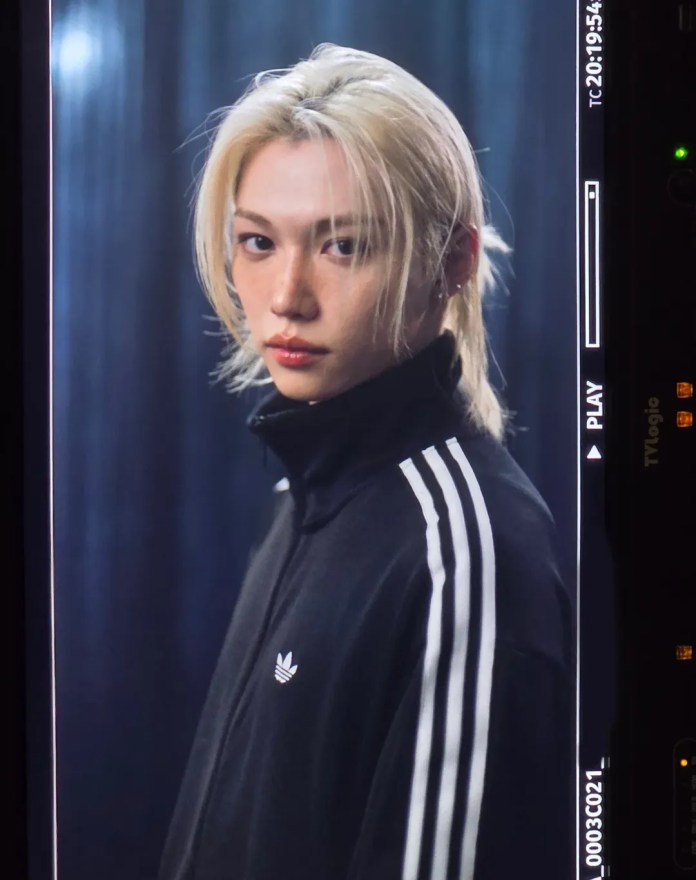 Close-up portrait of Stray Kids’ Felix with long blonde hair wearing a black Adidas jacket with white stripes, against a dark background.