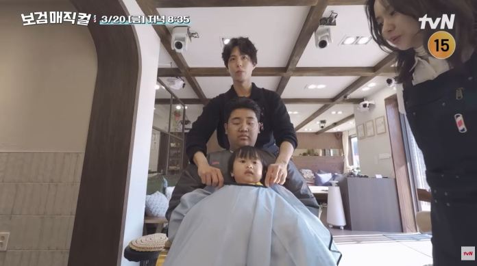 A child sitting in a hair salon chair covered with a hairdressing cape, being attended to by an adult stylist behind them while another adult adjusts the cape. Park Bogum stands nearby observing the scene.