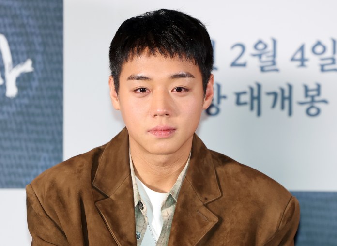 Park Ji-hoon with short hair, wearing a brown suede jacket and a light checkered shirt, looks directly into the camera at a promotional event.