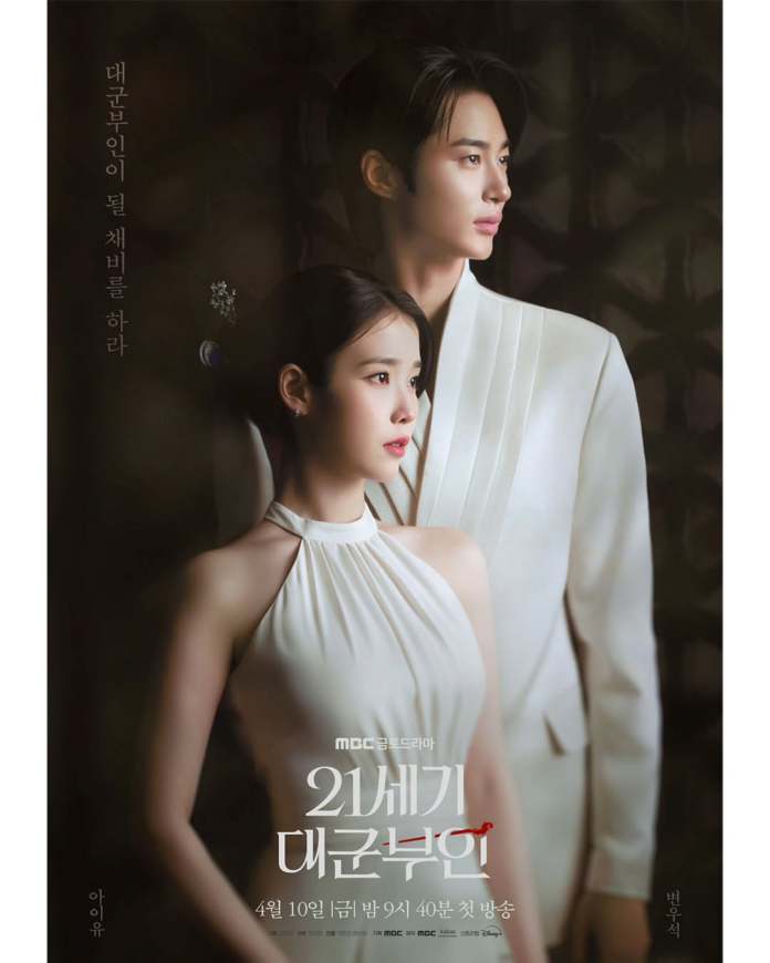 Promotional poster for a drama featuring IU and Byeon Woo-seok.I U in a white halter neck dress stands close to Byeon Woo-seok in a white suit, both looking serious and poised against a dark, textured background. Text in Korean is displayed on the side with the show's title and air date.