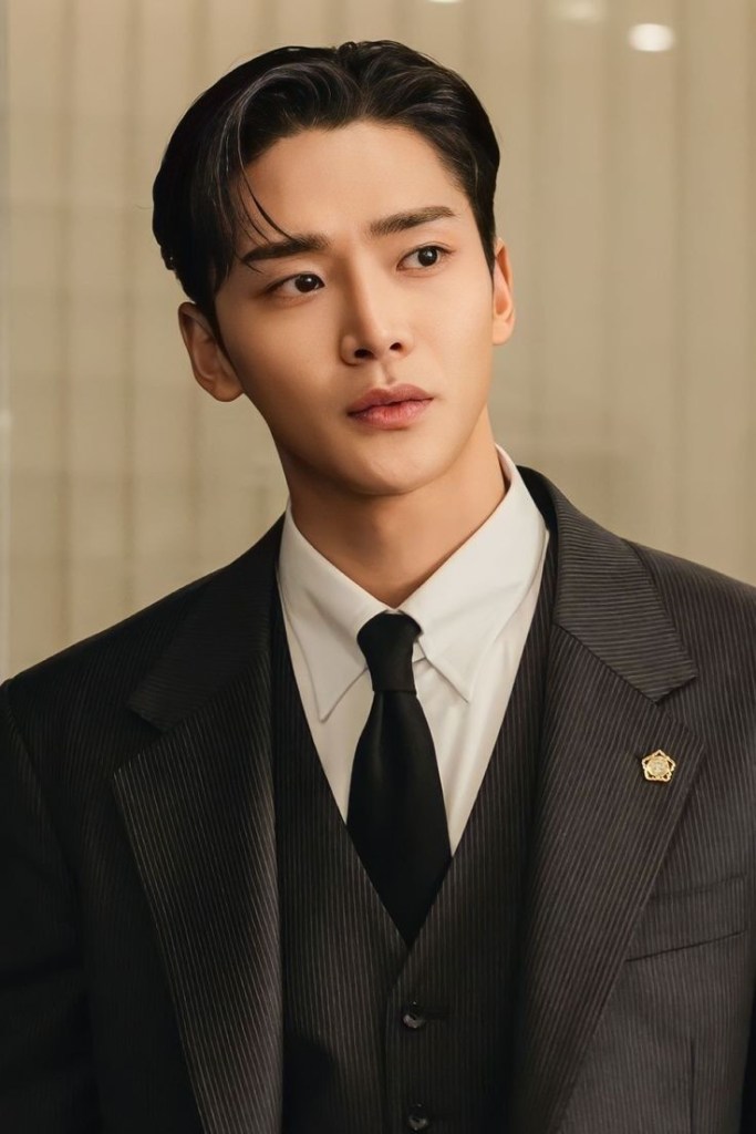 Close-up of Rowoon in a formal pinstriped suit with a tie, looking thoughtfully to the side against a blurred background.