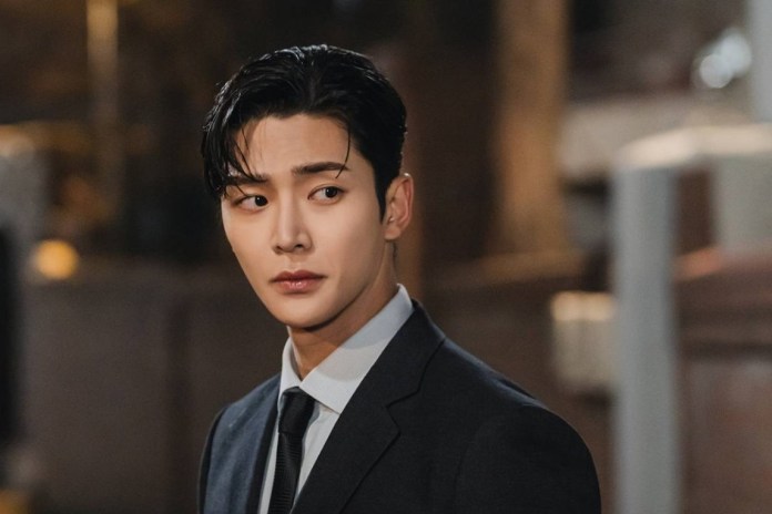 Rowoon with styled dark hair, wearing a formal suit and tie, looks thoughtfully to the side against a softly lit background, conveying a sense of intrigue.