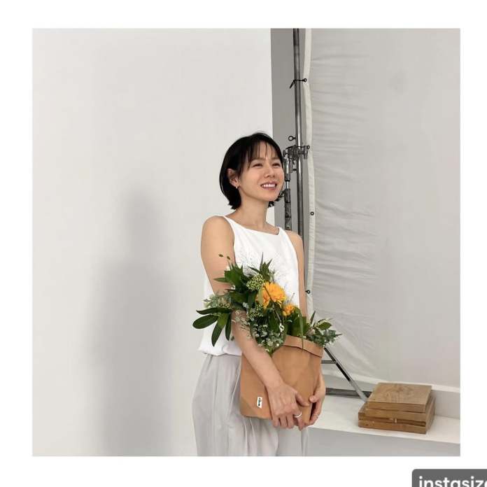 Son Ye jin smiling wearing a white tank top and light gray pants holds a bouquet of flowers in a tan bag, standing in a bright studio setting.