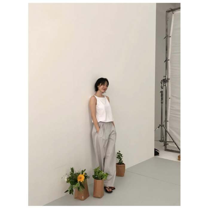 Son Ye jin in a white tank top and light gray pants stands against a plain wall, smiling casually. Two brown paper bags with plants and flowers are placed at her feet.