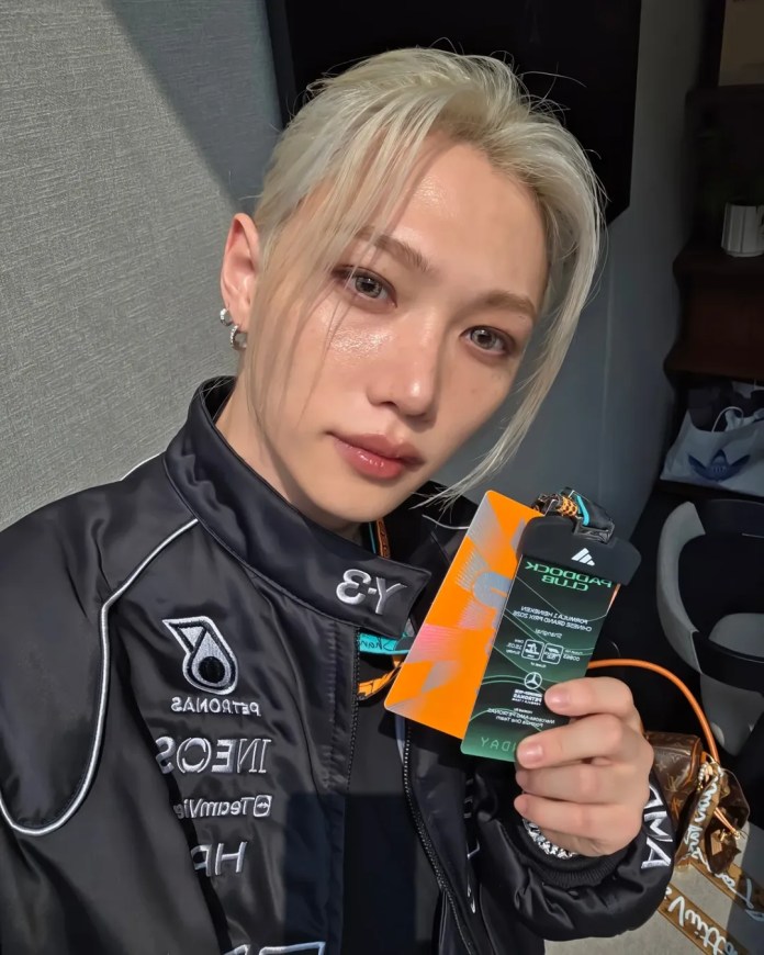 Stray Kids’ Felix with long, platinum blonde hair and a sleek hairstyle wearing a black jacket with branding. They are holding a colorful pass and smiling, set against a bright background.