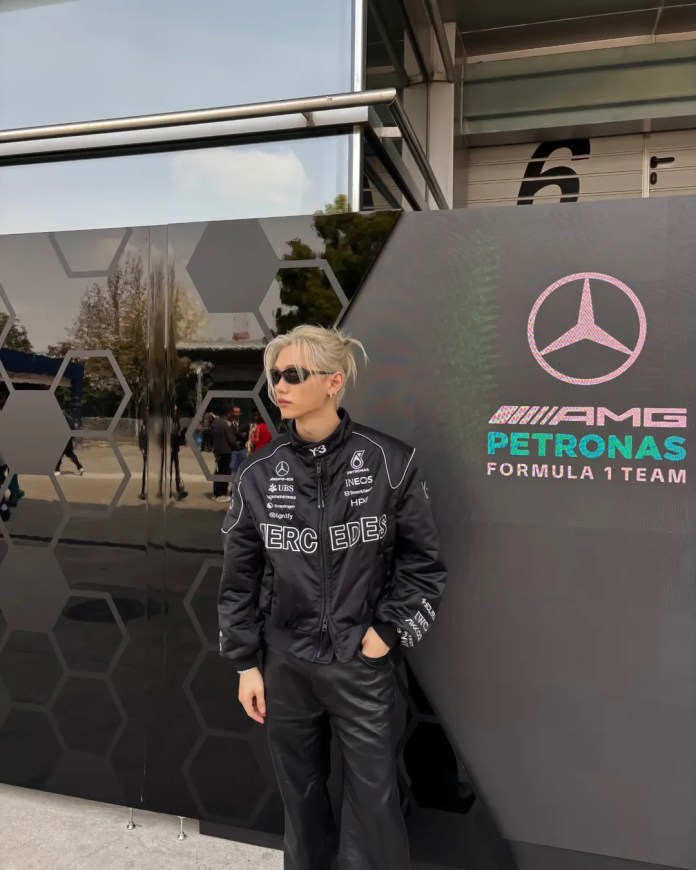 Stray Kids’ Felix wearing a black bomber jacket with Mercedes-Benz and Formula 1 team logos stands near a reflective wall featuring a hexagonal design and the AMG Petronas logo.