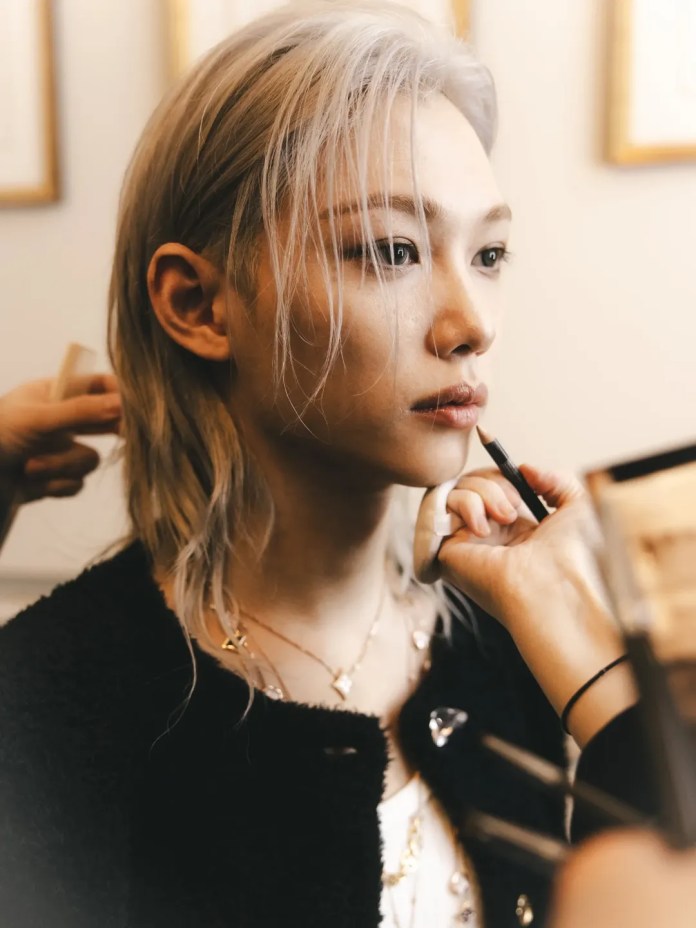 Felix with light-colored hair is having their makeup applied, focusing on the lips and eyes, in a well-lit room decorated with art.