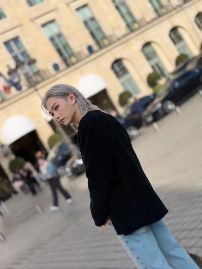 Felix with long, light-colored hair wearing a black jacket and light blue jeans poses in an urban setting with historic buildings in the background.
