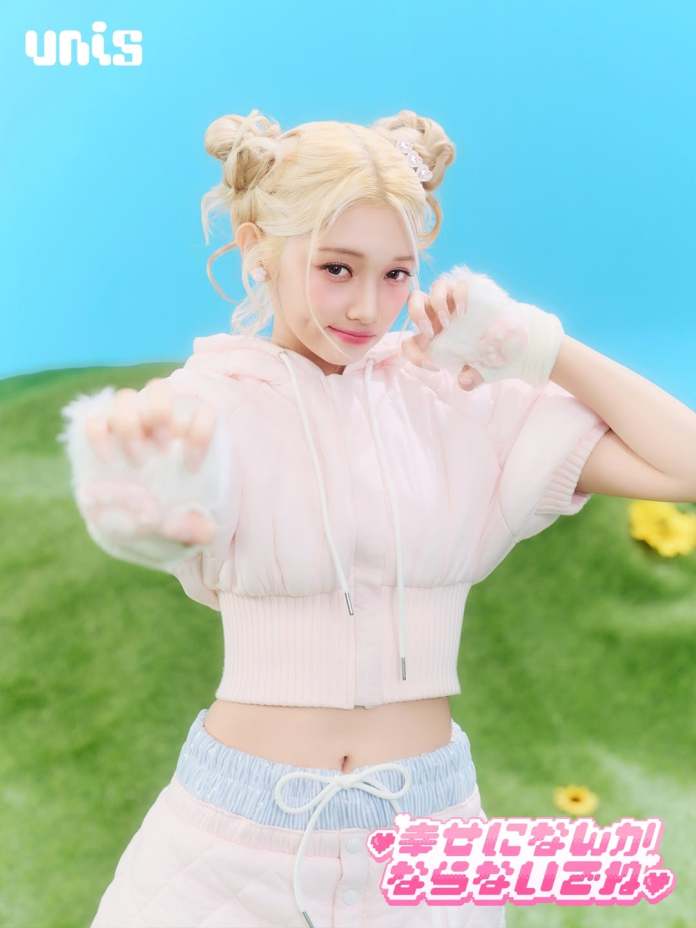 Gehlee with blonde hair styled in buns, wearing a pink cropped hoodie and light blue shorts, striking a playful pose against a bright blue background with green grass and flowers.