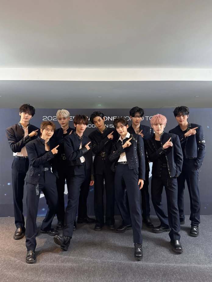 ZEROBASEONE posing together in stylish black outfits, all pointing in the same direction, with a simple backdrop featuring lights.