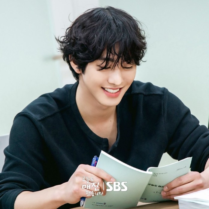 Ahn Hyo Seop with curly hair smiles while reading a script, seated at a desk with a pen in hand.