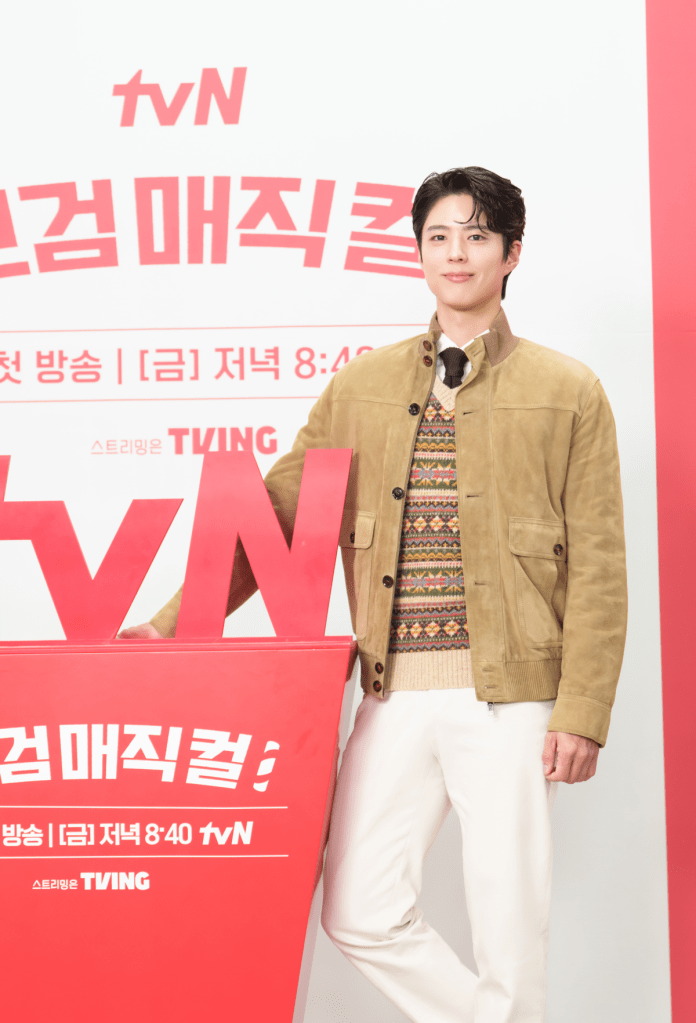 Park Bogum in a stylish outfit, featuring a beige jacket and patterned sweater, stands beside a large red backdrop displaying the tvN logo and promotional text.