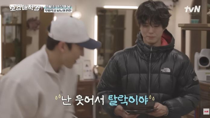 Park Bogum in a cozy indoor setting, one wearing a white jacket and baseball cap, and the other in a black puffer jacket. The scene contains playful interaction as one person appears to be laughing.