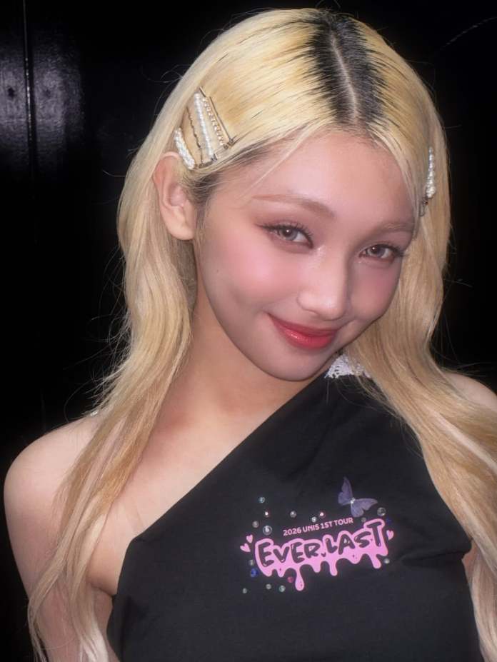 Gehlee with long blonde hair styled with hair clips, wearing a black top featuring a colorful tour logo, smiling at the camera.