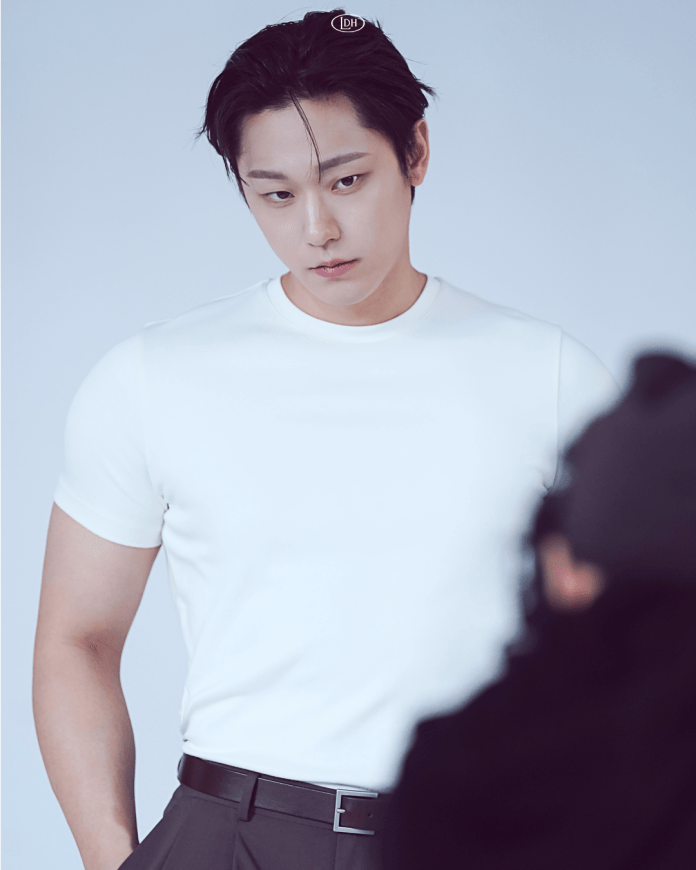 Lee Do hyun with short dark hair wearing a white t-shirt and black trousers, looking down thoughtfully against a plain background.