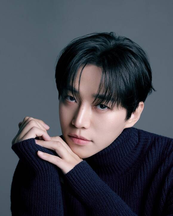 Portrait of a Lee Junho with dark hair, wearing a navy blue turtleneck sweater, gazing into the camera with a soft expression. The background is a neutral gray, enhancing the focus on the subject.