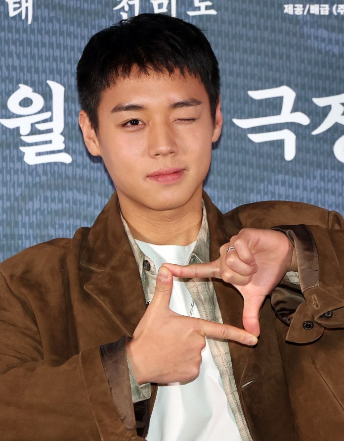 Park Ji-hoon with short, dark hair is winking and forming a frame with his hands against a patterned background.