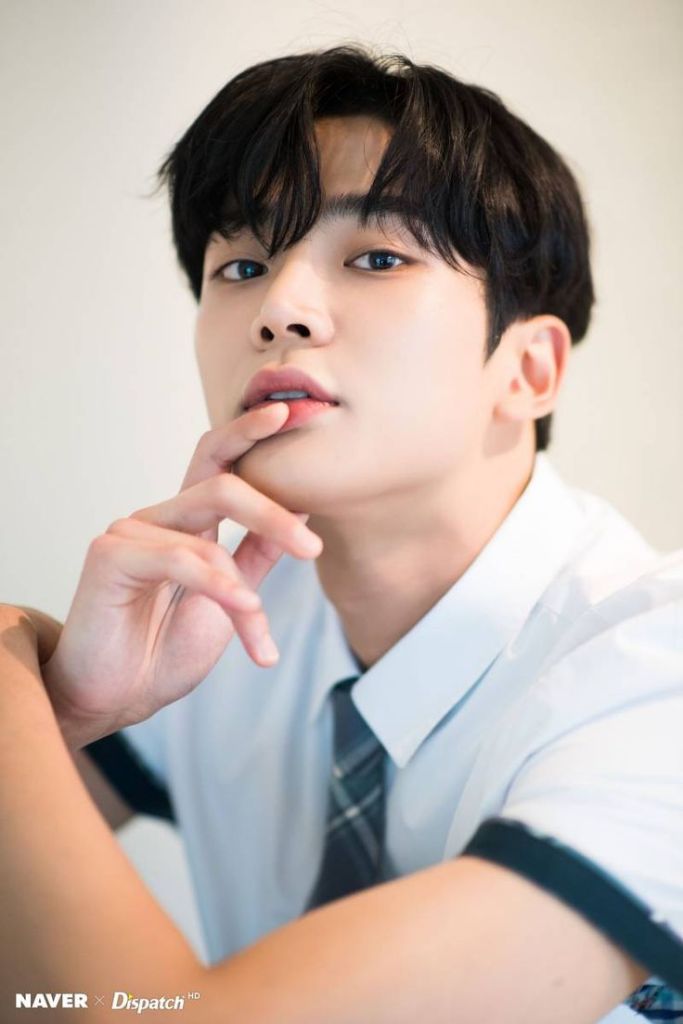 Rowoon with dark hair and blue eyes poses thoughtfully, wearing a white shirt with a plaid tie. He rests his finger on his lips, giving a contemplative look.