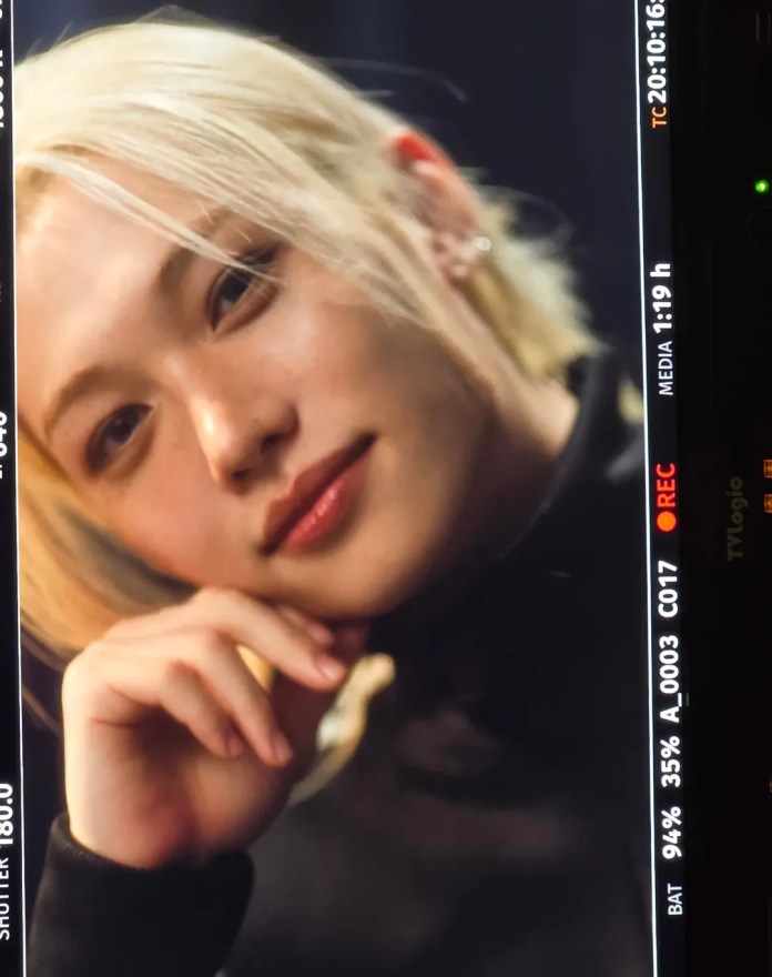 Close-up portrait of Stray Kids’ Felix with blonde hair, smiling slightly while resting their chin on their hand, wearing a dark outfit.