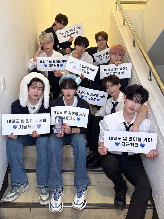 ZEROBASEONE sitting on stairs, each holding signs with Korean text that translates to 'I will know you tomorrow' in blue hearts. They have varied hairstyles and clothing styles, with a relaxed and friendly atmosphere.