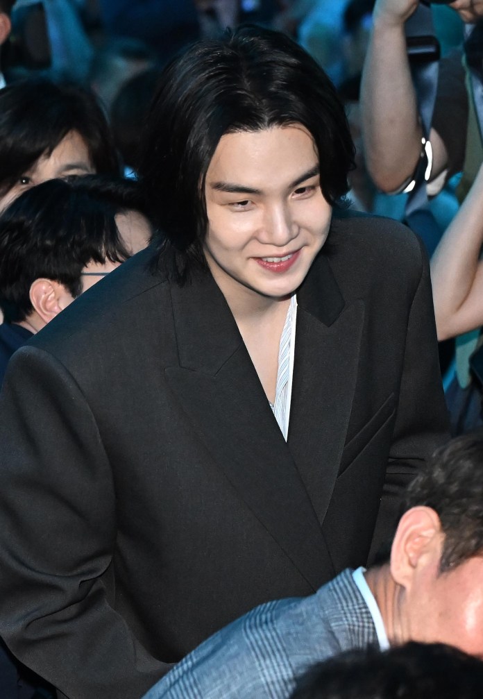 Suga with long dark hair and a warm smile, dressed in a black suit jacket, surrounded by a crowd.