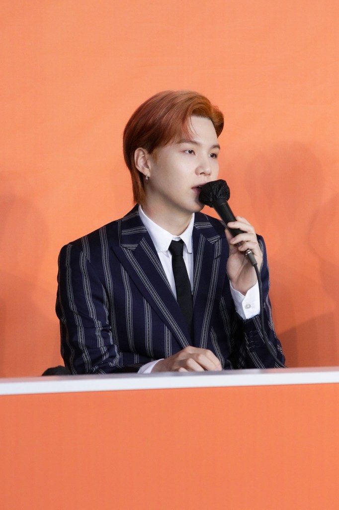 Suga with light brown hair and a striped suit is speaking into a microphone at a press event, with an orange background.