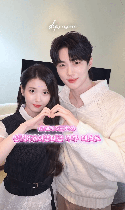 IU and Byeon Woo-seok form a heart shape with their hands while posing for a photo. They are smiling and dressed casually. The background appears to be a home setting.