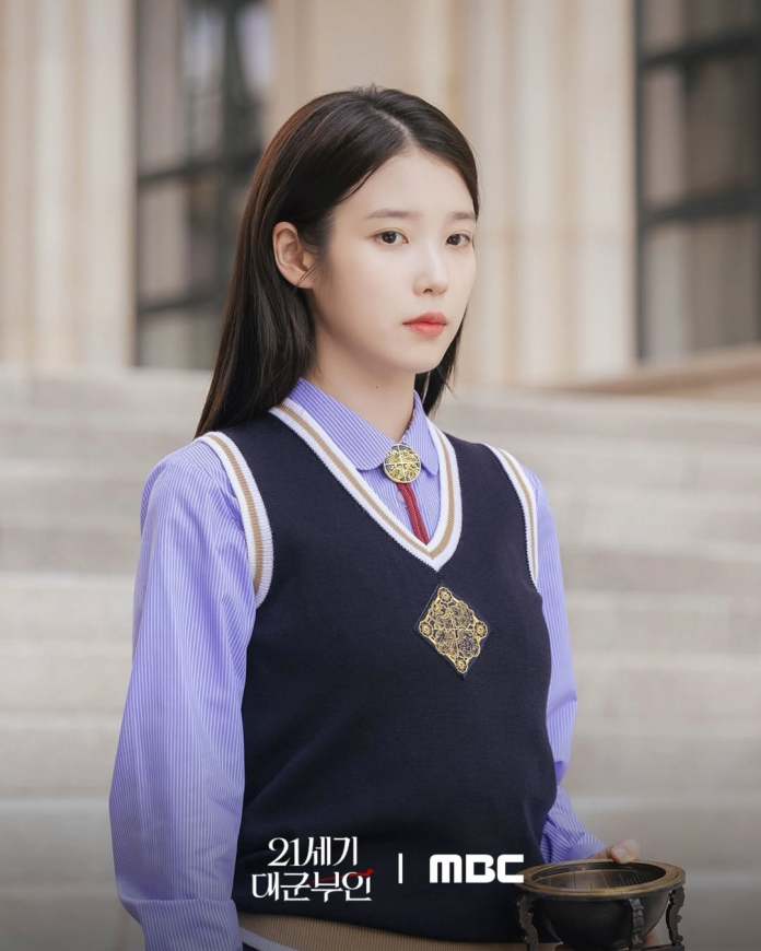 IU with long, dark hair is standing outdoors, wearing a navy blue vest over a light blue shirt. She holds a decorative bowl and looks serious, standing in front of stairs and a building.