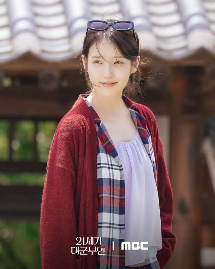 IU wearing a red cardigan over a plaid shirt, smiling softly in natural light with a traditional building in the background.