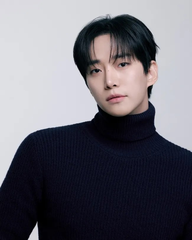 Lee Junho with short black hair and a turtleneck sweater poses against a light background, looking directly at the camera.