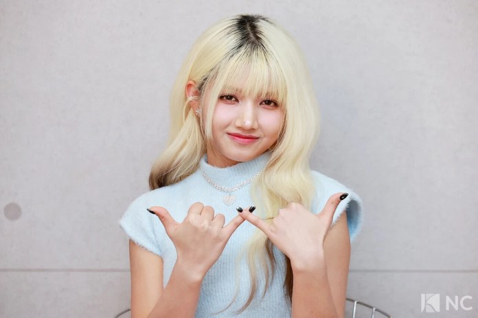 Hara with long, blonde hair wearing a light blue sleeveless top, making a hand gesture with both hands, smiling at the camera.