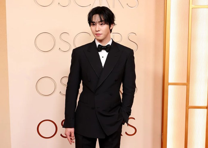Ahn Hyo Seop in a classic black tuxedo with a bow tie poses confidently on a red carpet backdrop featuring the word 'Oscars'.