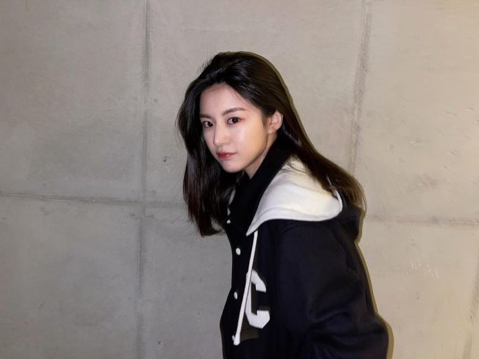 Go youn jung  with long dark hair is posing against a concrete wall, wearing a stylish black and white jacket.