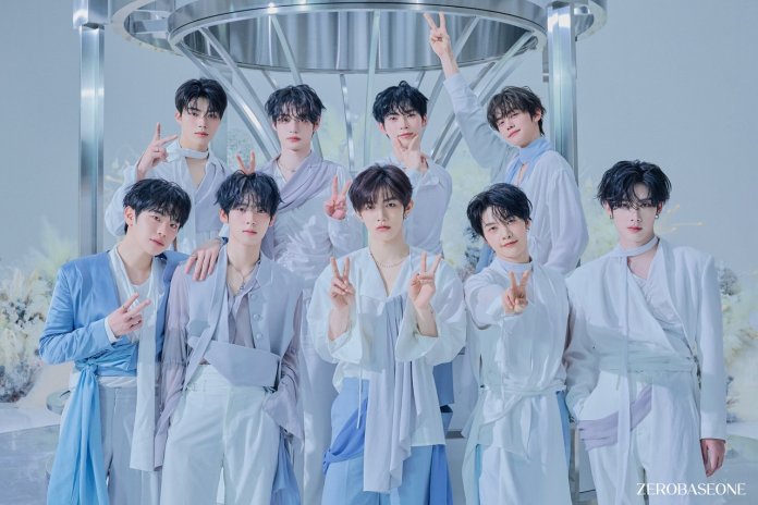 ZEROBASEONE posing together, all wearing stylish, light-colored outfits. They are standing in a semi-circle with some members making peace signs. The background features a futuristic design with soft lighting and decorative elements.