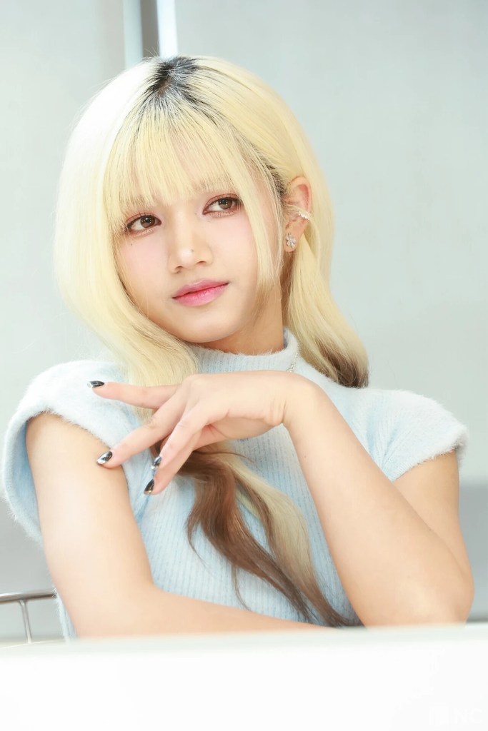 A close-up portrait of Hara with long, two-toned hair (blonde and brown) and a light blue sweater, posing thoughtfully with her hand near her face.