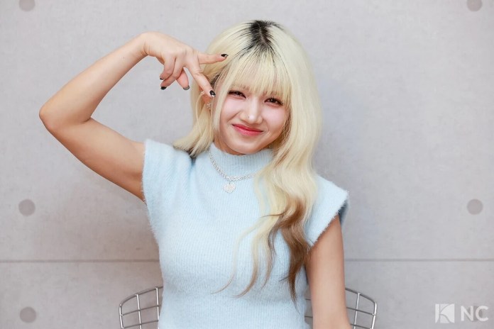 Hara with blonde hair styled in loose waves, wearing a light blue sleeveless top and a heart-shaped necklace, making a playful gesture with her hand.