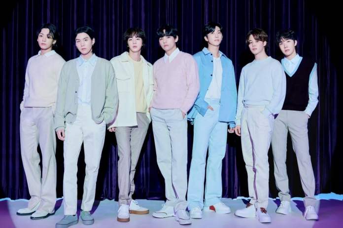 BTS standing in a line, dressed in stylish, pastel-colored outfits, against a dark purple backdrop.