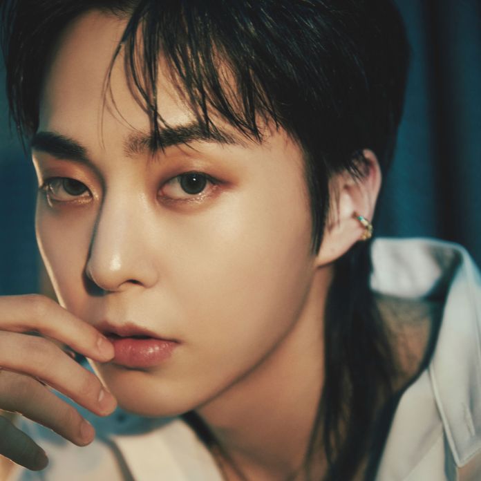 Close-up portrait of Xiumin with dark hair, wearing a white shirt, and holding their hand near their face, with a soft focus background.