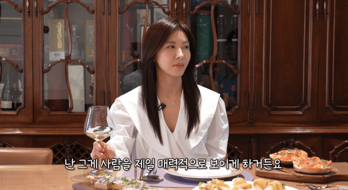Ha Ji-won sitting at a dining table holding a wine glass, with a spread of food in front of her, including appetizers and a wooden platter. She is dressed stylishly and appears to be in conversation.