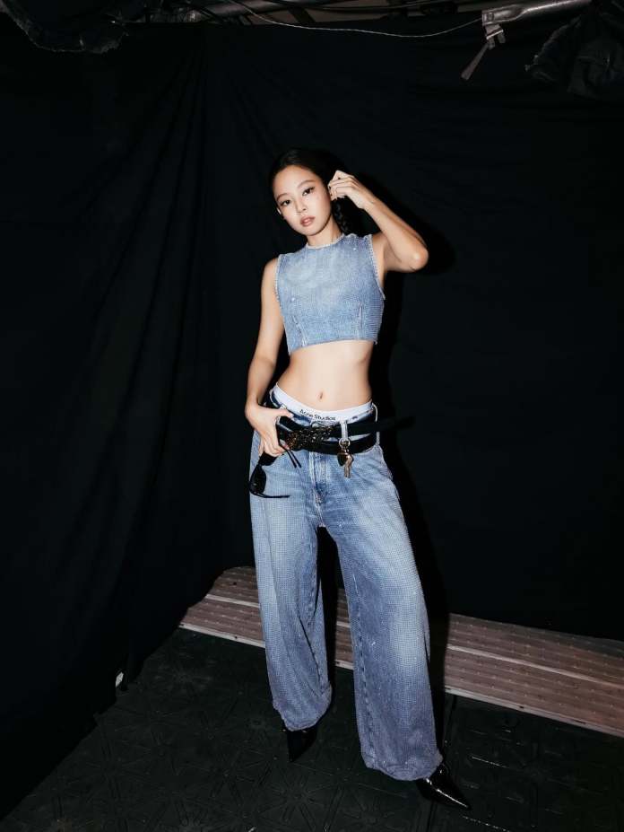Jennie posing in front of a black backdrop, wearing a denim crop top and loose-fitting denim pants, accessorized with a black belt and high heels.