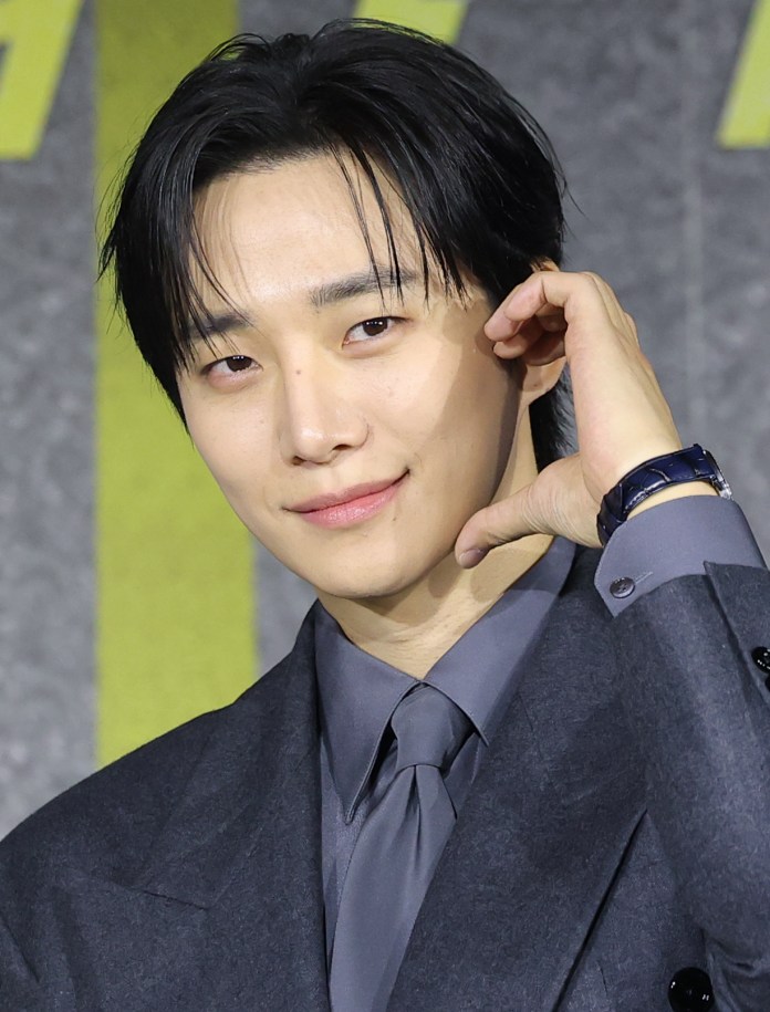 Junho in a gray suit and tie poses for the camera, making a hand gesture near his face against a textured background.