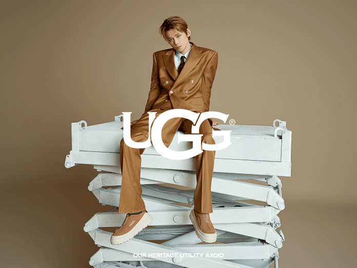 Yeonjun wearing a brown suit and casual shoes stands confidently on a stack of white furniture, with a neutral background and the UGG logo prominently displayed.