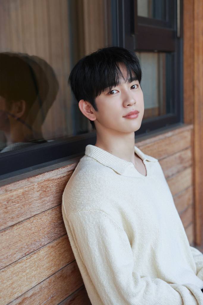 Park Jinyoung with dark hair wearing a light-colored sweater, sitting casually against a wooden wall and looking thoughtfully at the camera.