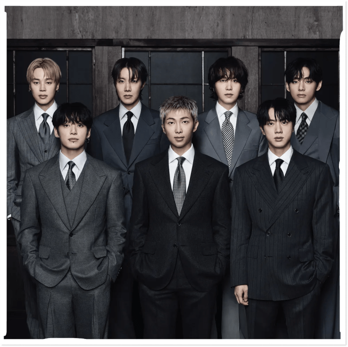 BTS wearing formal suits, standing side by side in a dimly lit setting. Each has a distinct hairstyle and facial expression, showcasing a blend of styles with gray and black suits.