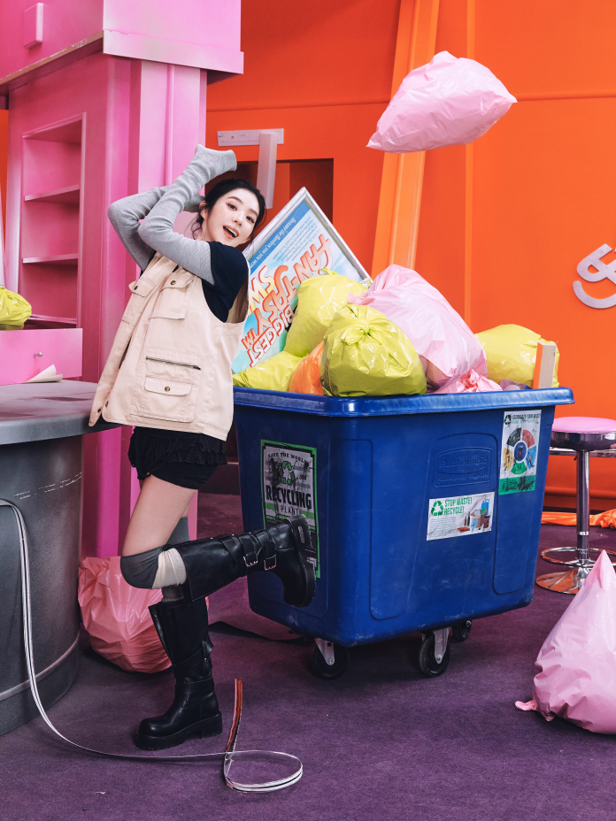 Irene playfully poses next to a blue recycling bin filled with brightly colored trash bags in a vibrant, colorful indoor setting.