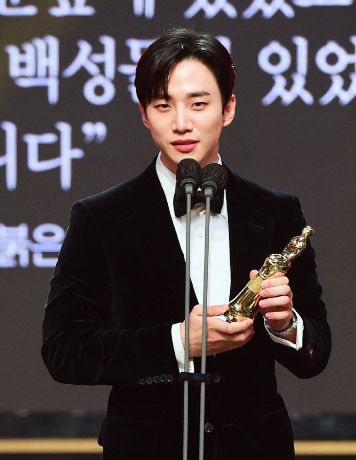 Junho in a black velvet tuxedo holding an award and speaking at a podium with microphones during an event, with text in the background.