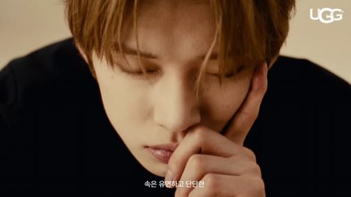 Close-up of a Yeonjun thoughtfully resting his chin on his hand, with his eyes closed, and a contemplative expression. The background is softly blurred.